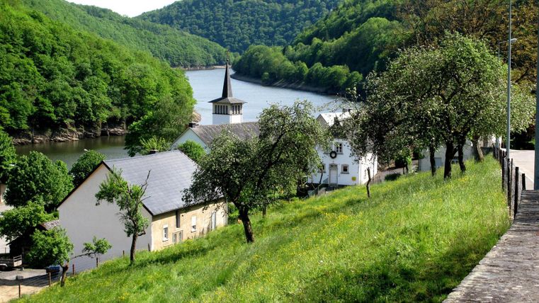Un paysage pittoresque avec une rivière, entourée de collines verdoyantes. Au premier plan se dressent des arbres et une petite agglomération avec un clocher.