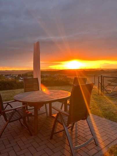 Een terras met een tafel en stoelen, dat bij zonsondergang is gefotografeerd. De warme kleuren van de zonsondergang verlichten het landschap op de achtergrond.
