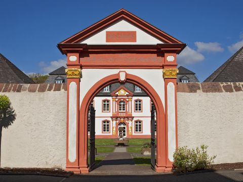 Een historisch gebouw met rode en witte muren is zichtbaar door een grote boog. De omgeving is groen en goed onderhouden, onder een heldere blauwe lucht.