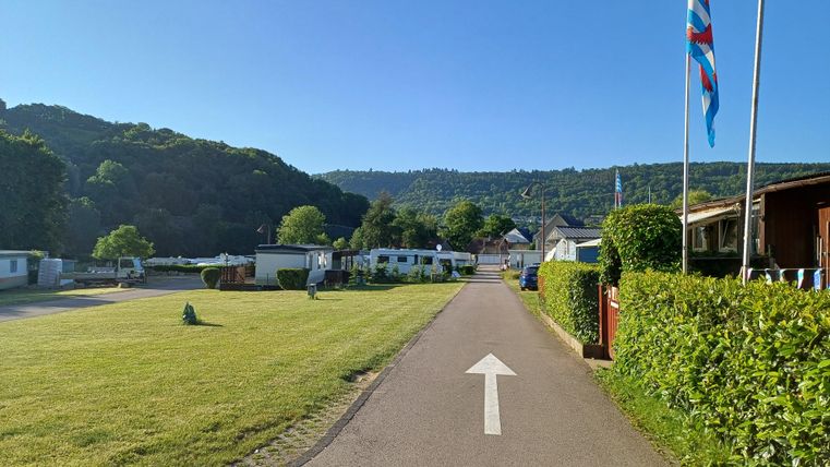 Een rustige camping met rechte paden en groene gazons. Op de achtergrond zijn bomen en heuvels te zien.