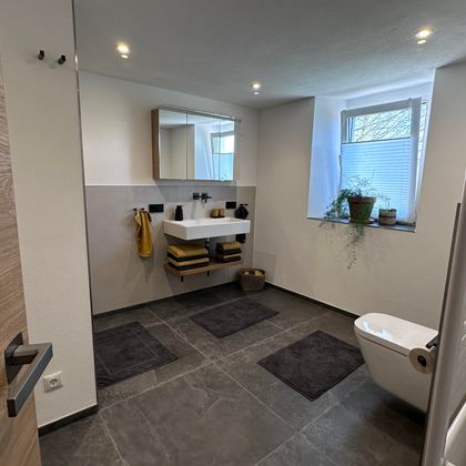 Ein modernes Badezimmer mit großen Fliesen und einem großen Spiegel. Es gibt Pflanzen auf der Fensterbank und eine helle, einladende Atmosphäre.