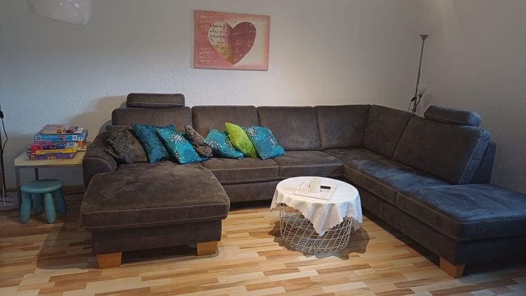 Ein gemütliches Wohnzimmer mit einem großen, grauen Sofa und bunten Kissen. Daneben steht ein Tisch mit einer Tischdecke und Bücher auf einem kleinen Tisch.