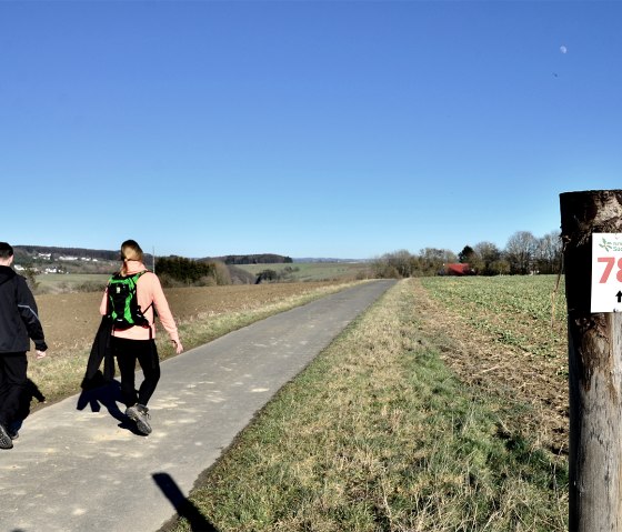 Zwei Wanderer auf einem Weg im Naturpark Südeifel. Ein Pfosten mit der Nummer 78 steht am Wegesrand. Der Himmel ist klar und blau., © TI Bitburger Land