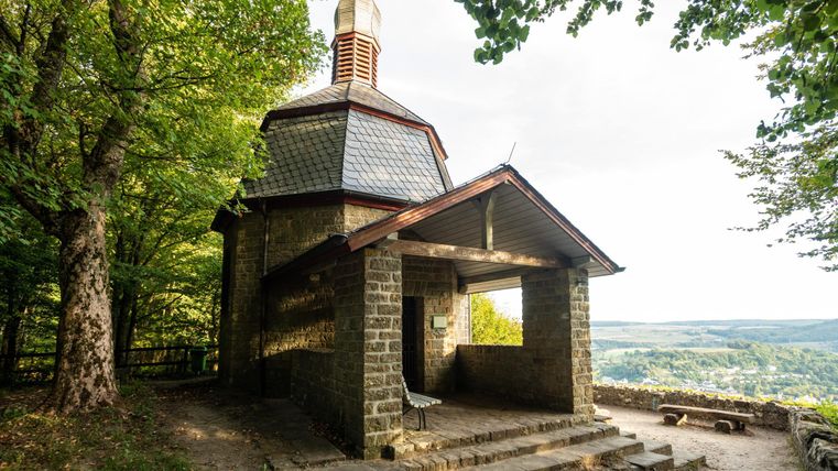 Eine kleine Kapelle aus Stein mit einem schlichten Dach, umgeben von Bäumen. Im Hintergrund erstreckt sich eine malerische Landschaft.