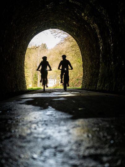 Zwei Radfahrer fahren durch einen dunklen Tunnel. Um sie herum ist ein schöner Wald und weiches Licht am Tunnelende.