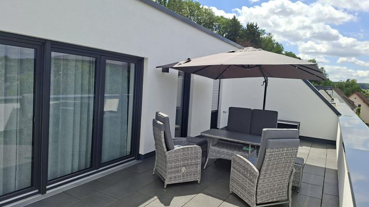 Une terrasse moderne avec une table à manger et des chaises confortables. Un parasol offre de l'ombre, tandis que la nature est visible en arrière-plan.