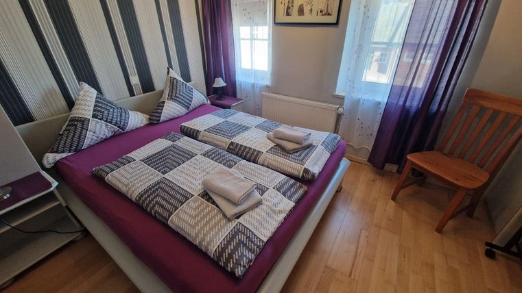 A cozy bedroom with a double bed and striped walls. Bright curtains let in plenty of light, and a wooden chair stands next to the bed.