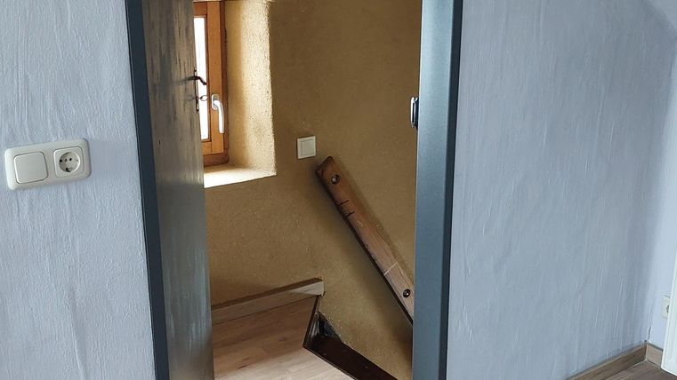 Een open deurframe met uitzicht op een trap en een klein raam. De muren zijn licht en de vloer is van hout.