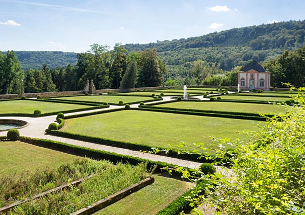 Französischer Garten mit geometrischen Hecken und Rasenflächen, im Hintergrund ein kleines Gebäude und bewaldete Hügel.