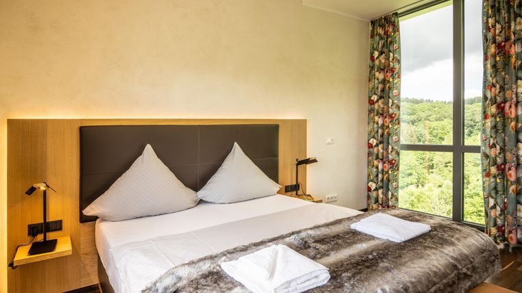 Une chambre moderne avec un grand lit et du linge de lit élégant. De grandes fenêtres donnant sur le paysage verdoyant laissent entrer beaucoup de lumière naturelle.