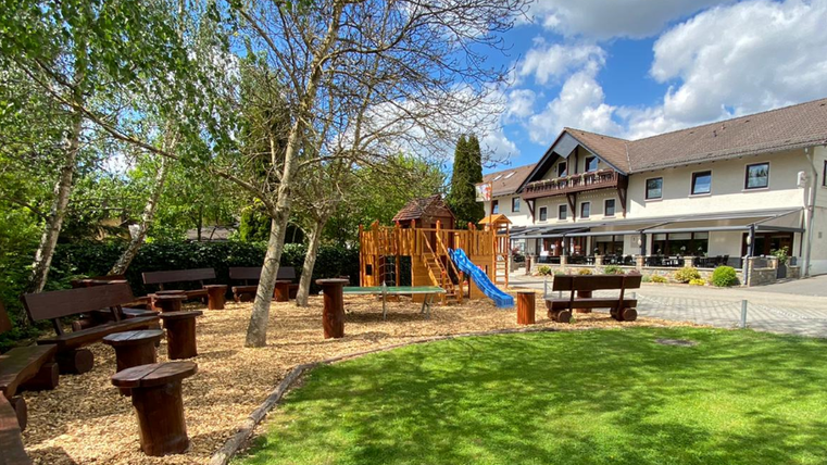 Een mooie buitenruimte met een speelhoek, houten zitplaatsen en een glijbaan. Op de achtergrond staat een gezellig gebouw met terras.