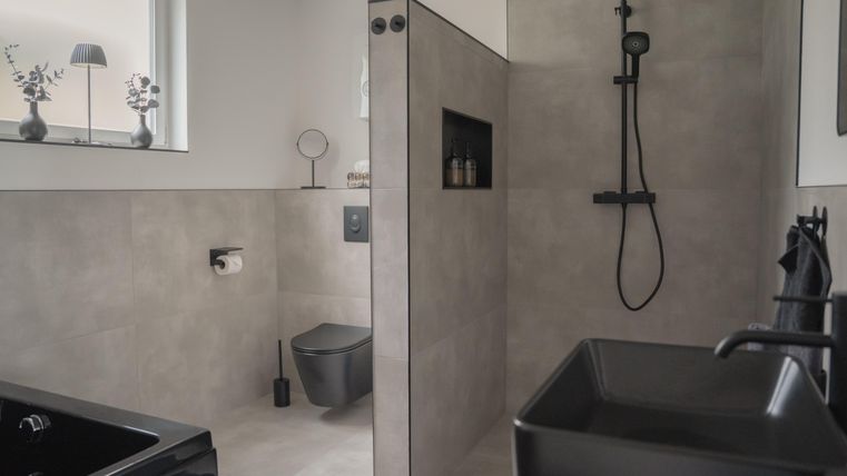 A modern bathroom with gray tiles and elegant black fixtures. The shower is open and well-lit, with a window that lets in natural light.