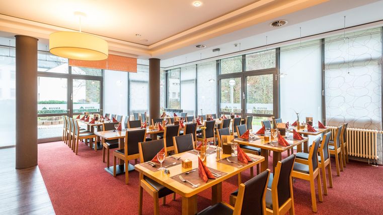 Een modern restaurant met elegante tafels en rode tafelkleden. Grote ramen zorgen voor veel daglicht en een aangename sfeer.