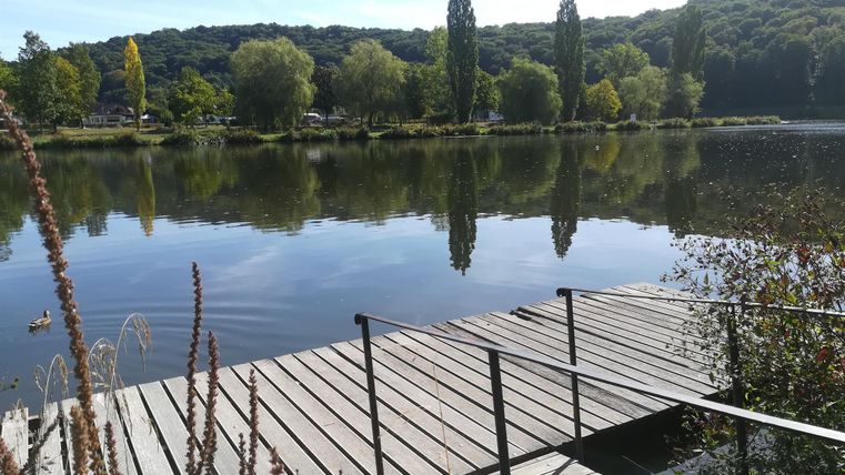 Ein ruhiger See mit einem Holzsteg und grünen Bäumen im Hintergrund. Das Wasser spiegelt die Umgebung und der Himmel ist klar.