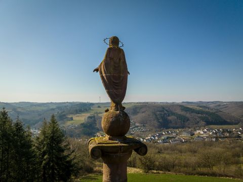 Eine Statue, die eine religiöse Figur darstellt, steht auf einer Säule und blickt über eine grüne Landschaft. Im Hintergrund sind Hügel und kleine Siedlungen zu sehen.