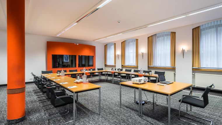 Een moderne vergaderzaal met meerdere tafels en stoelen. De muren zijn oranje en er zijn grote ramen met gordijnen.