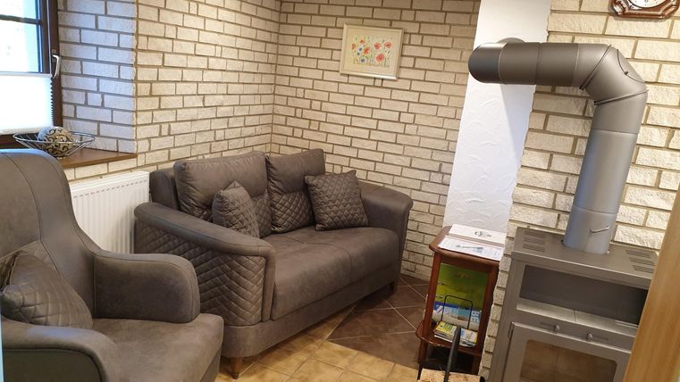 Un espace de vie confortable avec un canapé gris et un fauteuil. Les murs sont recouverts de briques et il y a une cheminée ainsi qu'une horloge au mur.
