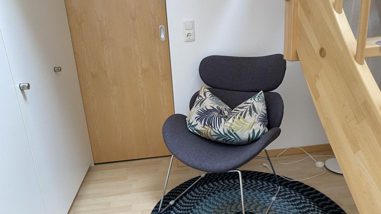 Ein gemütlicher Sessel mit einem bunten Kissen steht in einer Ecke eines hellen Raumes. Der Boden ist mit einem runden Teppich bedeckt, und eine Holztreppe führt nach oben.
