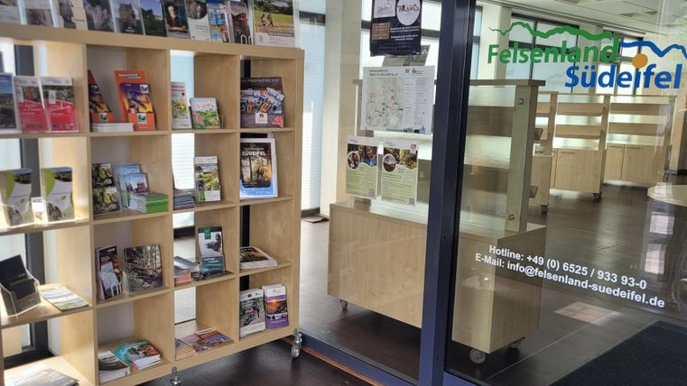 Ein Empfangsbereich mit einem Regal voller Prospekte und Informationen. Die Tür zum Büro zeigt die Website des Unternehmens "Felsenland SüdEifel".