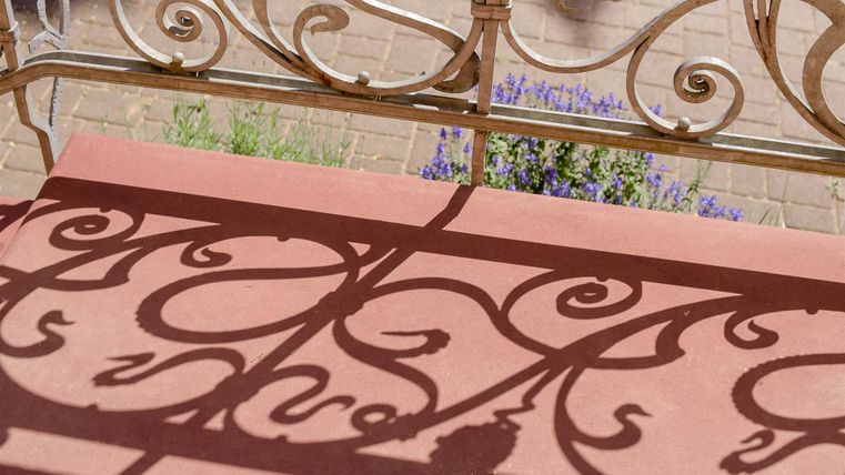Een decoratief ijzeren hek werpt fascinerende schaduwen op een rode oppervlakte. Op de achtergrond zijn paarse bloemen zichtbaar.