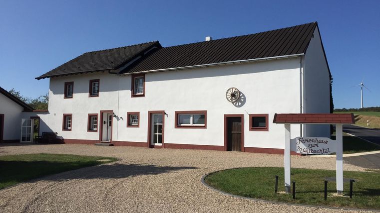 A large white house with a black roof and red window frames is situated in a quiet environment. The entrance area features a paved path and a small sign.