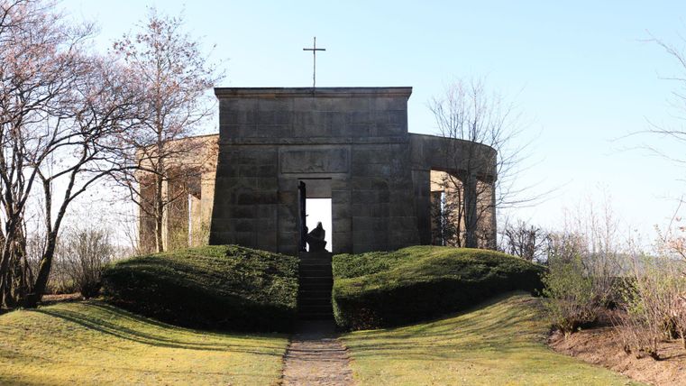 Ein historisches Bauwerk mit einem Kreuz auf dem Dach, umgeben von grünen Hügellandschaften. Die klare, sonnige Atmosphäre schafft eine ruhige Stimmung.
