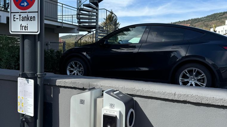Ein schwarzes Elektroauto steht neben einer Ladestation. Im Hintergrund sind ein modernes Gebäude und eine Wendeltreppe sichtbar.