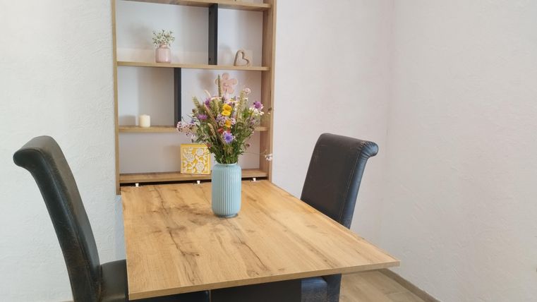 Een gezellige eetruimte met een houten tafel en twee zwarte stoelen. Op de tafel staat een vaas met kleurrijke bloemen.