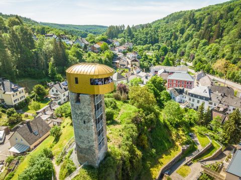 Ein historischer Turm mit einem modernen, goldenen Dach steht über einer malerischen Stadt. Im Hintergrund erstrecken sich grüne Hügel und Wälder.