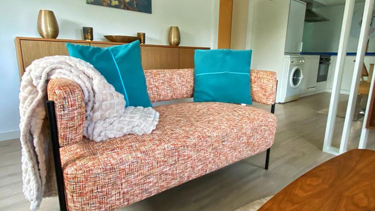 Ein gemütliches Sofa mit buntem Stoff und dekorativen Kissen steht in einem modernen Raum. Im Hintergrund sind einige Möbel und ein Waschraum zu sehen.