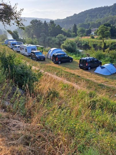Un camping en pleine nature avec plusieurs camping-cars et tentes. En arrière-plan, on voit des collines douces et un ciel clair.