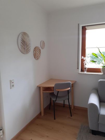 Une pièce lumineuse avec une petite table et une chaise dans le coin. Des feuilles décoratives sont accrochées au mur, et une fenêtre avec des plantes permet une lumière naturelle.