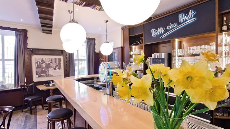 A stylish bar with a round counter and bright wood. Fresh yellow flowers are placed on the bar countertop, while the decor appears cozy.