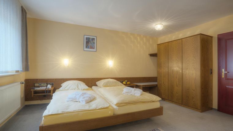 Ein einfaches Hotelzimmer mit zwei Einzelbetten und einem Holzkasten. Die Wände sind in einem warmen Gelbton gestrichen, und es gibt ein Fenster mit Tageslicht.