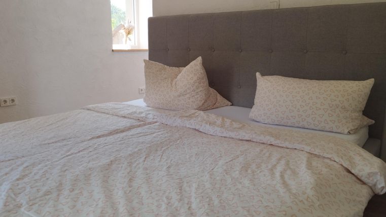 Een net bed met een grijze hoofdeinde en blauwe kussens. Het beddengoed heeft een subtiel patroon en de ramen laten natuurlijk licht binnen.