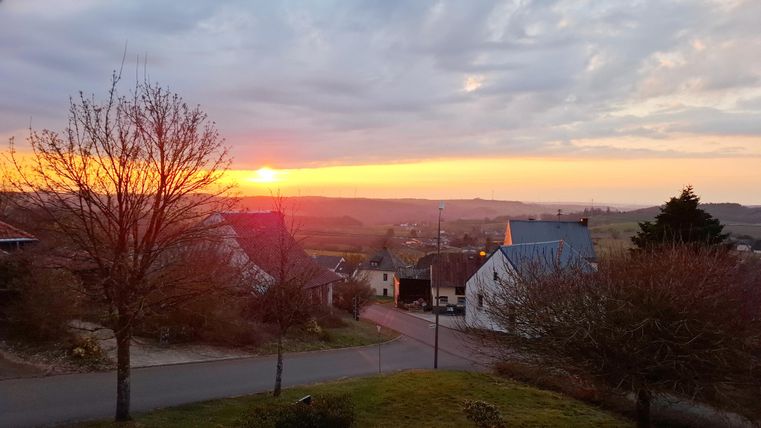 Ein malerischer Sonnenuntergang über einer ruhigen Landschaft. Häuser und Bäume rahmen die Szene ein, während der Himmel in warmen Farben leuchtet.