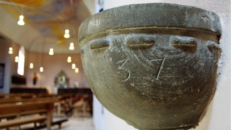 Un bol en pierre avec le chiffre 37 est fixé au mur d'une église. En arrière-plan, des bancs d'église et un éclairage doux sont visibles.