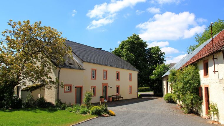 Ein idyllisches Anwesen mit einem großzügigen, grünen Garten. Im Vordergrund sind mehrere Häuser und ein Kiesweg zu sehen, unter einem klaren blauen Himmel.