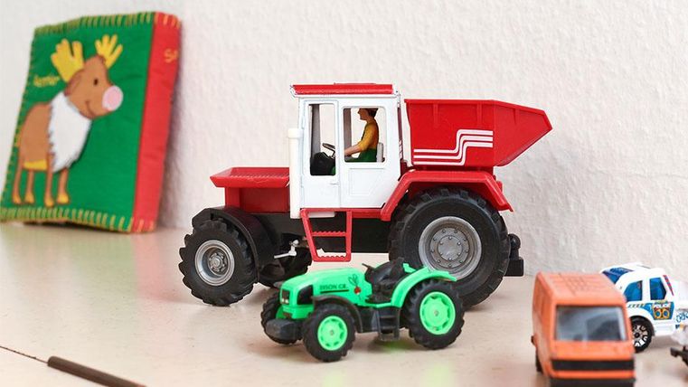 Ein roter Spieltraktor steht neben einem grünen Spielauto und einem kleinen orangen Fahrzeug. Im Hintergrund ist ein Kinderbuch mit einem Tiermotiv zu sehen.