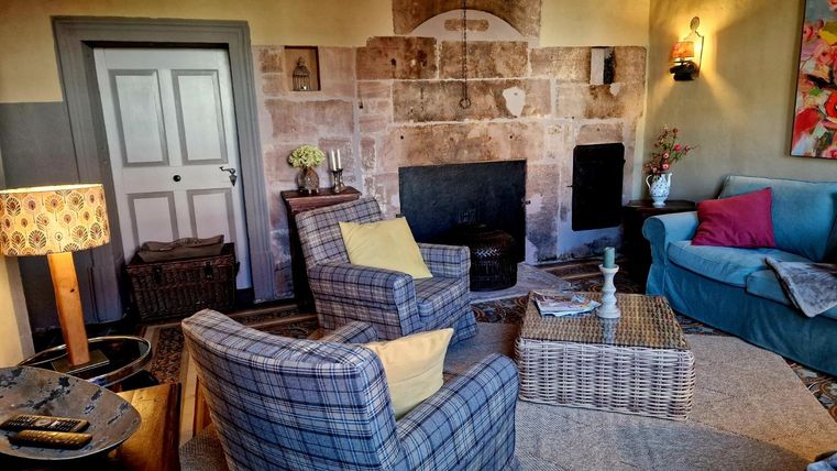 Een gezellige woonkamer met een open haard, twee fauteuils en een bank. De muren zijn in warme kleuren geschilderd en er zijn decoratieve elementen zoals bloemen en een tapijt.