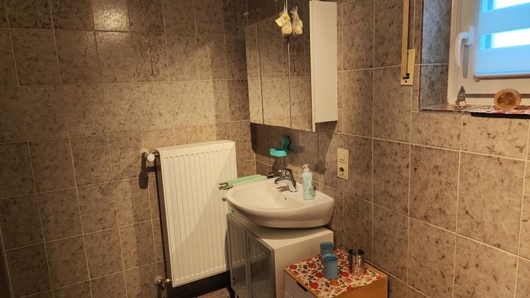 Ein kleines Badezimmer mit grauen Fliesen und einem Waschbecken. Die Einrichtung umfasst einen Spiegel und einen Heizkörper.
