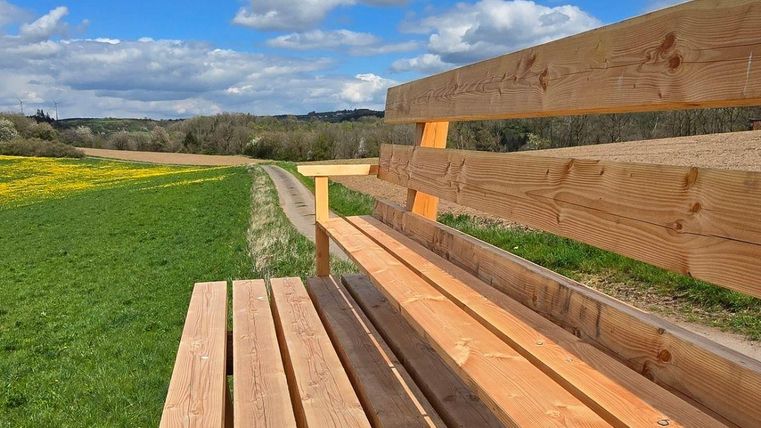Een houten bank staat op een groen veld onder een blauwe lucht met wolken. Op de achtergrond zijn weiden en bomen te zien.