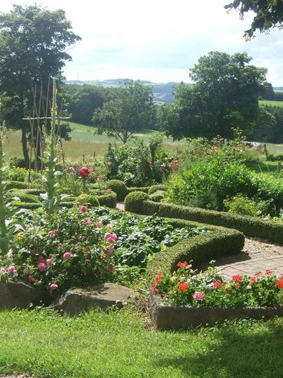 Ein schöner Garten mit bunten Blumen und gut gepflegten Hecken. Im Hintergrund sind sanfte Hügel und Bäume zu sehen.