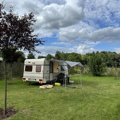 Une caravane est posée sur une pelouse verte, entourée d'arbres et de buissons. Le ciel est partiellement nuageux et la scène semble paisible et accueillante.