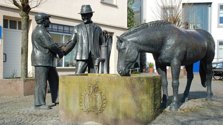 Eine bronzene Statue zeigt zwei Männer, die sich die Hand geben, neben einem Pferd an einem Brunnen. Die Szenerie ist städtisch und vermittelt ein Gefühl von Gemeinschaft.