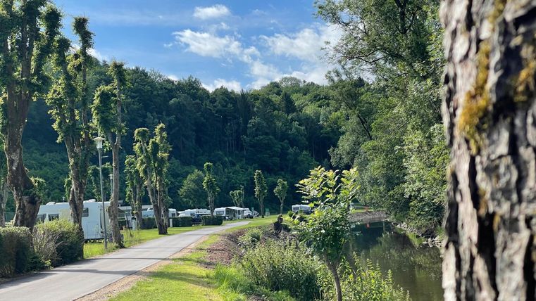 Ein ruhiger Campingplatz am Fluss, umgeben von Bäumen und grüner Natur. Der Himmel ist klar mit wenigen Wolken.
