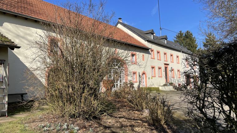 Une maison ancienne avec des cadres de fenêtres rouges et un jardin. Les environs sont verts et ensoleillés.