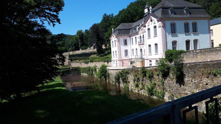 Un bâtiment historique se dresse au bord d'une eau calme. Entourée d'arbres et d'herbe verte, la scène dégage une atmosphère de tranquillité et de paix.