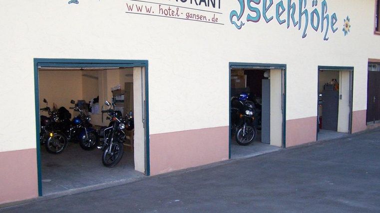 Eine Motorradgarage mit mehreren Motorrädern steht im Vordergrund. Darüber befindet sich das Schild eines Restaurants namens "Isekkhöhe".