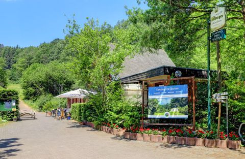 A cozy restaurant in the midst of nature with green trees and colorful flowers. The path leads to the entrance area, where tables are set up outside.